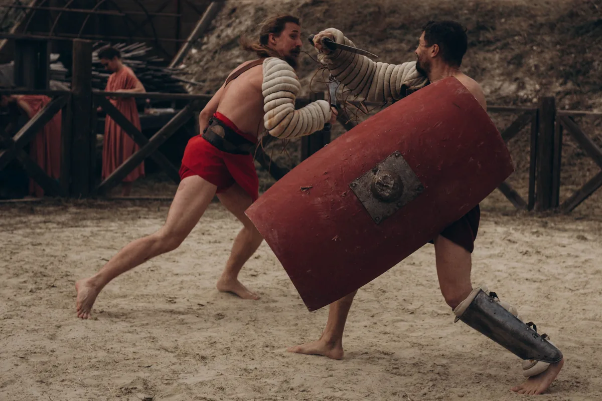 Two gladiator re-enactors fight in a sand arena, one lunging with a dagger over a large red shield while the other twists away