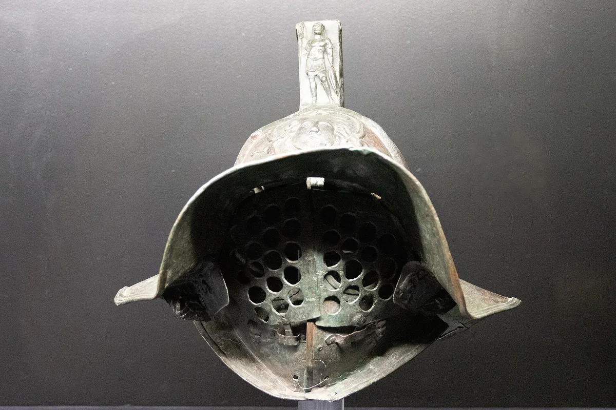 A bronze gladiator's helmet of the murmillo type with a broad brim, a grilled visor and a ridged crest, displayed on a stand