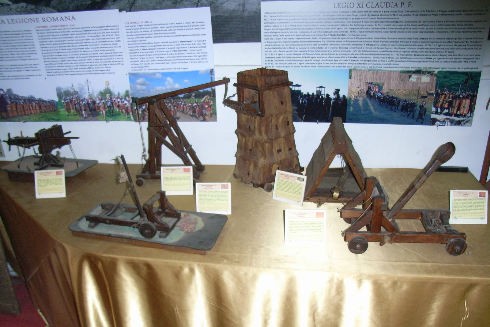 A table of wooden models of Roman siege engines, including a catapult, a ballista and a siege tower, with printed labels beside each and panels about the Roman legion on the wall behind
